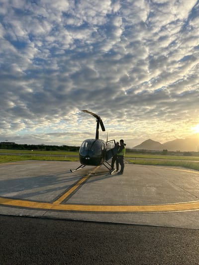 The image depicts a helicopter on a helipad during either sunrise or sunset, characterized by strong backlighting from the low sun on the right horizon, casting long shadows. Two adult males are present near the dark-colored Robinson R44 helicopter, which is stationary on the concrete helipad marked with yellow lines. One male, wearing dark clothing, is partially inside the helicopter's cockpit, with the door open. The second male, wearing a dark top and a bright yellow-green high-visibility vest, stands outside the open door, facing towards the helicopter.
The environment comprises a concrete helipad transitioning to an asphalt foreground, bordered by green grass. Beyond the airfield, a dense line of green vegetation is visible, followed by several undulating mountains in the background, characteristic of the landscape in Rio de Janeiro, Brazil. A construction site with a crane is partially visible on an elevated area on the far left. The sky is partially covered with scattered cumulus or stratocumulus clouds, illuminated by the golden light of the low sun on the right, contrasting with cooler blue and grey tones elsewhere. The scene's specific mountainous backdrop points to a location within the metropolitan area of Rio de Janeiro, likely an airfield such as Jacarepaguá.