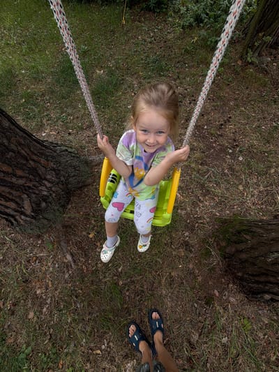 A young girl with blonde hair is sitting on a yellow swing in a wooded area. She is wearing a colorful t-shirt and leggings with heart and rainbow designs. She is smiling and looking at the camera. The swing is attached to ropes that are hanging from a tree. The ground is covered in grass, leaves, and pinecones. The perspective of the shot is from someone standing in front of the swing, looking up at the girl. Their feet, wearing dark sandals, are visible at the bottom of the frame.