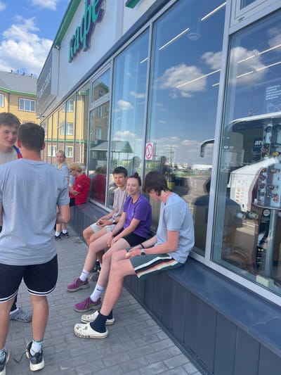 The image depicts a group of teenagers standing in front of a building in Kazan, Russia. They are close to a storefront with a glass display window, which shows advertisements for eyeglasses and sunglasses. The reflection in the glass reveals a bright, sunny day with blue skies and clouds.
The teenagers are dressed in casual clothing, including t-shirts, shorts, and sneakers. Some are wearing socks and sandals. They appear to be looking at something inside the store. A few of them are standing close together.
The background shows a building with yellow and green accents. The overall setting suggests a typical urban environment on a sunny day in Kazan, Russia.