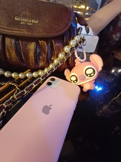 A close-up shot shows a glossy, dark red handbag with the embossed text "MIKAXIQI & BAG EST. 1985" in gold lettering. A decorative chain with faux pearls and a darker chain is attached to the handbag, leading to a pink Stitch-like keychain. To the right of the handbag and chain, a pink iPhone case is visible, displaying the Apple logo. A person's arm in a patterned maroon top is partially visible in the upper right corner. The objects are placed on a dark, reflective surface, possibly a table. The background is blurred, but hints of illuminated signage, possibly from a restaurant or shop, are present. There is no indication of the location being in Nematjan, Uzbekistan.