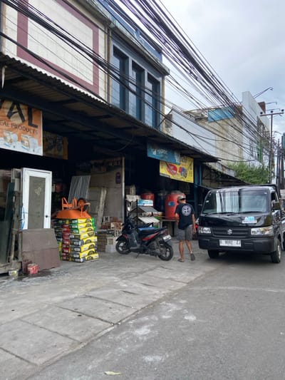 A man in a black t-shirt and shorts is walking on the sidewalk in front of a building. The building has a large sign that says "REAL PRODUCTS." There are several stacks of building materials on the sidewalk in front of the building, including a stack of bricks, a stack of cement bags, and a wheelbarrow. There is a black motorcycle parked next to the stack of cement bags. There is a black pickup truck parked in the middle of the road in front of the building. The truck is a Suzuki Carry. The truck's license plate is D 8082 ZJ. The image was taken in the city of Kecamatan Margahayu, Indonesia, at the coordinates -6.97 latitude and 107.574 longitude. The sky is overcast and there are many power lines in the image.  The building facade is primarily made of brick. The sidewalk is made of concrete and is uneven in places. The road is asphalt and appears to be a narrow street.