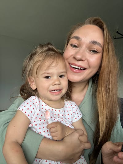 The media file is a close-up, low-angle selfie of a woman holding a young child. The woman, with long blonde hair, is smiling broadly, showing her teeth. She is wearing a green top. The child, facing the camera, also has a wide smile and is wearing a white top with red polka dots. The child's right hand is visible, holding a small white measuring spoon with a pink substance, possibly medicine or food. They are positioned closely together, suggesting an affectionate moment. The background is a plain, muted green wall, with a hint of a ceiling fixture at the top left corner. The lighting is soft and appears to be natural, likely from a window, suggesting it could be daytime. The overall emotion conveyed is happiness and warmth between the two individuals. There is no visible text in the image. The location context provided is Kharnavy, Russia.