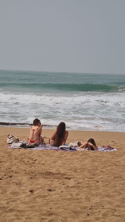 The image shows three women on a beach in Nahariyya, Israel. Two are sitting up and one is lying down. They are all wearing swimwear. The beach is sandy and there are waves breaking in the background. There are towels and bags around the women. The sky is overcast.