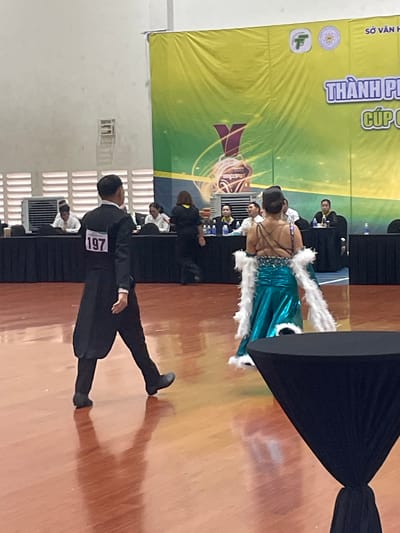 The image shows a ballroom dancing competition or performance at an indoor venue. In the foreground, a male dancer in a black tailcoat with a white bib displaying the number "197" is walking away from the camera. To his right, a female dancer in a shimmering turquoise gown with white feather accents on her arms is also walking away from the camera. In the background, a large green and yellow banner is visible with Vietnamese text and logos. Several people are seated at tables behind the dancers, possibly judges or spectators. The floor is a polished wooden surface. This event is likely taking place in Thuận An, Vietnam, as indicated by the banner which appears to be from the Department of Culture and Information of Thuận An City.