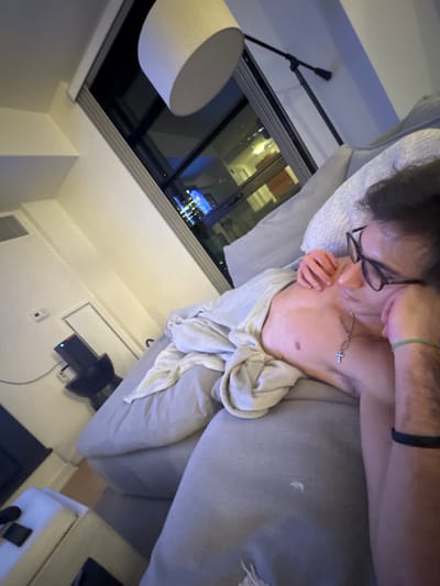 The image displays an interior scene at night, likely within a high-rise residential unit or hotel room located in Boston, United States. Through a large, floor-to-ceiling window occupying the right side of the frame, a nighttime city skyline is visible, featuring various illuminated buildings and a prominent skyscraper lit in blue.
A male individual is positioned on his back on a light-grey sectional sofa, partially covered by a light-colored blanket or sheet over his lower torso and chest. He is shirtless, wears black-rimmed glasses, and has a silver chain with a cross pendant around his neck. His right arm is bent, with his hand near his face. His left wrist displays both a green and a black bracelet.
To the left of the sofa, a dark, cylindrical side table supports a black, rectangular electronic device exhibiting a green-red indicator light on its top surface. Above this table, on a white wall, a white rectangular panel and a small white wall-mounted control unit are visible. In the upper-center of the image, a floor lamp with a white drum shade extends upward. In the bottom-left corner, a white upholstered ottoman holds a black smartphone. The room features white walls and is dimly lit by interior artificial light, supplemented by the ambient city lights from outside the window.