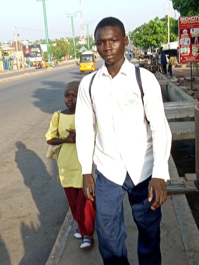 This media file captures two individuals walking along a paved road in Tumfure, Nigeria. The main subject is a young man in the foreground, facing the camera. He is wearing a white long-sleeved collared shirt with a school emblem on the left breast, and dark blue trousers. A backpack is slung over his right shoulder. To his left and slightly behind him, a younger child, appearing to be a girl, walks in the same direction. She is wearing a bright yellow long-sleeved garment and dark red trousers. She carries a patterned bag on her left side.
The scene appears to be a busy street with a roadside environment. In the background, there are buildings, trees, and streetlights. A yellow three-wheeled vehicle, likely a motorized rickshaw or tuk-tuk, is visible on the road. A large billboard advertising "RICH CITY POPCORNS" with images of popcorn and ice cream is prominently displayed on the right side of the image, near the road. The presence of the billboard and the type of vehicles suggest an urban or semi-urban setting in Nigeria.
The lighting indicates that it is daytime, likely late afternoon, given the long shadows cast by the individuals on the road. The weather appears to be clear and sunny. The expressions on the faces of the subjects are neutral. The overall atmosphere seems casual, possibly depicting a daily commute or a walk.
