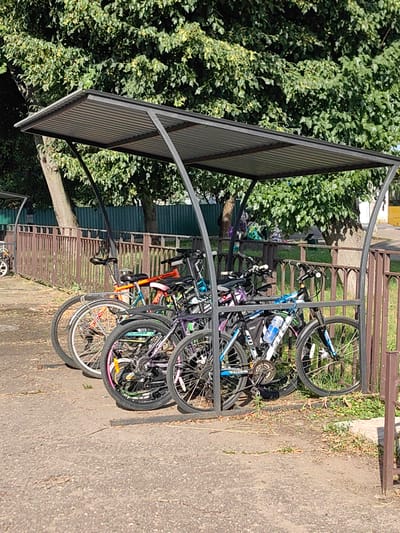 This is a vertical, medium-shot photograph taken on a sunny day in Fanipal, Belarus. The image depicts a row of bicycles parked under a metal canopy designed to shelter them from the elements. The canopy has a corrugated metal roof and dark grey metal support posts.
The bicycles are of various colors and designs, including a bright orange and blue bike, a purple and black bike, and a black and blue bike with a water bottle mounted on the frame. Some of the bicycles have colorful accents on their rims. They are parked closely together, suggesting a communal bike rack.
In the background, a rusty brown metal fence runs horizontally across the frame. Beyond the fence, lush green trees with dense foliage dominate the upper portion of the image, casting dappled shadows. The ground beneath the canopy and in front of the fence is a mix of asphalt and dry, patchy grass with fallen leaves. The overall scene appears to be an outdoor area, likely in a residential or public space, designed for bicycle parking. The bright sunlight indicates it is daytime. There is no visible text in the image.