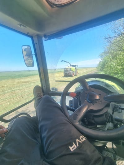 The image depicts a view from inside the cab of a tractor. The driver is visible, wearing dark pants with the word "NOVA" printed on the right thigh, and a tan work boot on the right foot. The tractor's steering wheel is prominently in the foreground, with other controls and the dashboard visible. Through the front windshield, another tractor is seen operating in a field of recently cut grass. The sky is blue with some clouds visible near the horizon and through the windshield. Based on the mention of the location, this scene is likely in or near Svoboda, Bulgaria, but there are no specific visual markers to confirm this directly. The scene suggests agricultural activity, specifically harvesting or field maintenance.