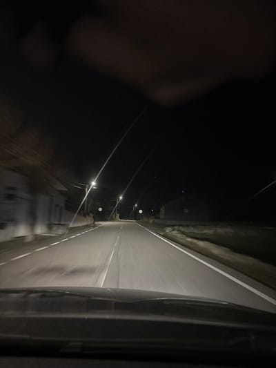 A nighttime, forward-facing view from inside a moving vehicle on an asphalt road in Carrazeda de Ansiães, Portugal, is depicted. The vehicle's dark dashboard or hood occupies the lower foreground. The two-lane road, illuminated by the vehicle's headlights and multiple streetlights, features white solid shoulder lines and a broken white center line. Streetlights extend into the distance, generating visible lens flares, indicating artificial illumination of the scene. The left side of the road presents blurred outlines of structures consistent with rural or residential buildings, accompanied by utility poles and two circular road signs with indiscernible markings due to distance and low light. The right side of the road reveals open, dark terrain, possibly fields, extending towards a large, unlit building with a barn-like or industrial silhouette in the mid-ground. The sky above is dark and cloudless. No persons are present in the scene.
