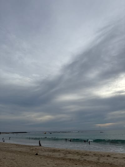 The image depicts a beach scene at what appears to be Meunasahbale, Indonesia. The sky is overcast with dramatic grey clouds, with a sliver of brighter sky visible on the right horizon. The ocean is a turquoise color with moderate waves, and several people are visible in the water, some appearing to be surfing or playing. There is a yellow inflatable object in the water, and a small dark boat is further out at sea. On the sandy beach in the foreground, a few individuals are present, and some debris, possibly a coconut, is on the sand. The overall impression is a somewhat moody, but active, beach day.