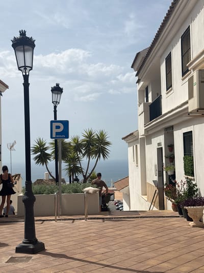 This is an outdoor, eye-level, medium shot of a street scene in Benalmádena, Spain. The image is captured on a sunny day with a partly cloudy sky. The foreground is dominated by a textured reddish-brown tiled pathway, casting a long shadow from a tall, black, ornate lamppost on the left.

In the mid-ground, a white building with white stucco walls and a red-tiled roof stands on the right, featuring barred windows and balconies. Next to it, a pathway descends, with another building on the left that is partially visible. Between the buildings, a few palm trees are seen, along with some shrubbery.

On the left side of the pathway, a woman in a black dress walks away from the camera. Further back, another person, possibly a child, is seated. In the center of the frame, a blue parking sign for bicycles is mounted on a pole next to a lamppost. A man is pushing a cart on the pathway behind the sign.

In the background, the blue sea is visible, with some rooftops and other buildings interspersed. The overall scene depicts a charming street in a Spanish coastal town.