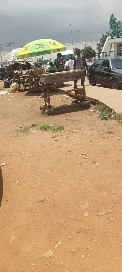 The image captures an outdoor scene in Kuru Dungus, Nigeria, likely a marketplace or a roadside vendor area. In the foreground, a rough wooden table supported by rocks sits on the dusty, reddish-brown ground. Scattered around the table are various items, including plastic bags, debris, and some green grass peeking through the dirt. A large, open green umbrella with "GLO VOICE & DATA PLANS" and "GLO LTE ADVANCED" written on it is positioned behind the table, providing shade.
Several people are visible in the background. One man wearing a grey tank top with the number 9 on it is standing and looking towards the camera, smiling. Another person in a colorful floral shirt is standing next to him, with a child in a green shirt nearby. A white van is parked behind them, and a black car is visible on the right side of the frame, driving on a paved surface. Further in the background, other stalls and vehicles suggest a busy commercial area. Overhead, a cloudy sky looms. A large map-like structure with green and white coloration is visible in the far background on the right.