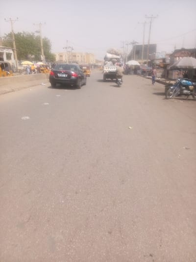 The image displays a daytime street scene in Dakata, Nigeria, characterized by a hazy atmospheric condition that softens distant features. The central element is a granular, light brown-grey asphalt road extending into the background, appearing to be a multi-lane thoroughfare. Small, scattered white debris is visible on the road surface in the foreground and midground.
In the midground, traffic proceeds away from the viewer. A dark-colored Honda Civic sedan with a rear license plate (partially legible as "YBC 9XX") occupies the left lane. In the right lane, a white Toyota pickup truck, heavily laden with large, irregularly shaped bundles wrapped in light-colored material, is visible. A person on a small motorcycle follows directly behind the pickup truck.
To the left of the road, a concrete median separates the main carriageway from a roadside area. This area contains multiple yellow motorized rickshaws/tricycles and several pedestrians. Beyond this, white-walled buildings are visible, interspersed with green foliage from trees, and utility poles supporting power lines extend along the street.
To the right of the road, a series of market stalls or shops line the thoroughfare, featuring various coverings including white and striped umbrellas or canopies. Several individuals are present in this area, some walking. A light blue motorcycle is parked on its kickstand on the right shoulder. Similar to the left side, utility poles with power lines and additional buildings are discernible in the background. The scene depicts a busy urban commercial environment.