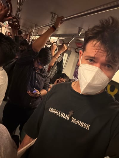 In the dimly lit interior of a crowded Delhi Metro train car, a man in the foreground wearing a white face mask and a black t-shirt with white Cyrillic lettering, appears to be taking a selfie. The t-shirt reads "СВОБОДНЫЙ ЧЕЛОВЕК" (Svobodnyy chelovek), which translates to "Free person" in Russian. A wooden cross necklace is visible around his neck. His blue eyes are visible above the mask. Behind him, other passengers are holding onto overhead rails or are looking at their phones. One man in a striped dark shirt and glasses is looking down at his phone. The train car's ceiling is lined with metal poles and handrails, characteristic of public transportation in Delhi. The overall atmosphere suggests a busy commute during peak hours.