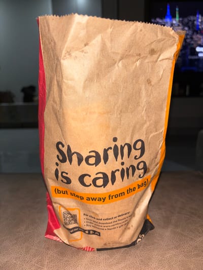 A slightly crumpled brown paper bag is positioned on a surface, likely a couch. The bag's text reads "Sharing is caring (but step away from the bag)". Additional text on the bag provides information about clicking and collecting or delivery: "Scan and download the Nando's app. Order online www.nandos.co.za. Buy someone a Nando's gift card." A QR code is also visible.
The paper bag appears to be from Nando's. This is inferred from the logo and address (Nando's, South Africa) on the bag. Sandton, South Africa is not evident in the image. The background includes a blurred TV or screen, and a dark cabinet or wall. The image is well-lit, but with shadows.