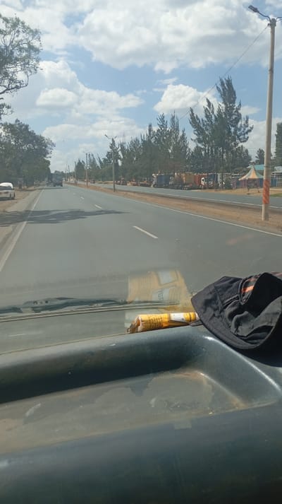 A sunny day scene from inside a vehicle, overlooking a straight road. The road has white lane markers and trees line the right side with utility poles. In the distance, parked trucks and a tent structure are visible. On the vehicle's dashboard, there is a rolled-up object wrapped in yellow packaging and a black hat. The dashboard is dark green and worn. The sky is blue with scattered clouds.