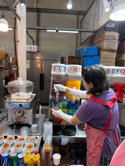 In an indoor market setting, likely in Jeju City, South Korea, a woman wearing a purple t-shirt and a red and white striped apron stands behind a counter filled with drinks and snacks. She is wearing white disposable gloves and appears to be dispensing a beverage from a large dispenser labeled "포도" (grape) into a clear plastic cup. Next to this dispenser is another one filled with bright orange liquid, labeled "오렌지" (orange). To the right of the orange juice dispenser, there is a third dispenser with red liquid.

On the counter in front of the woman are several small white cups filled with bite-sized fried food, possibly chicken. Various plastic cups and straws are neatly stacked to the right. Cans of soda and bottles of yellow and red sauces are also visible on the counter. Behind the beverage dispensers, a large silver refrigerator is present. The environment appears to be a food stall or a small shop within a larger market, with overhead lighting and shelving units visible in the background.