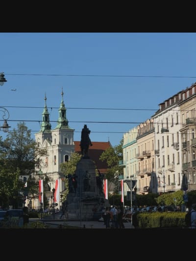 The image shows a street scene with buildings, a monument, and people. In the background, there are several multi-story buildings with balconies and windows. One building on the left features two green-domed towers. In the foreground, a large stone monument with statues stands in the center. Several people are visible walking and standing near the monument and on the sidewalks. The sky is clear and blue, and there are trees and streetlights present. The scene appears to be a public square or plaza in a European city, likely during the daytime. The presence of Polish flags suggests a connection to Poland, not Bucharest, Romania.