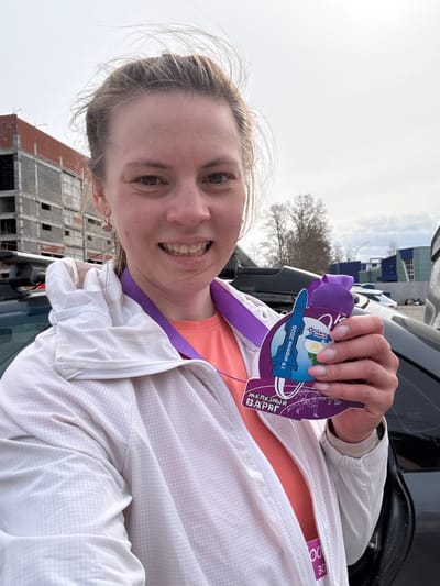 A smiling woman holds a medal outside in Neftekamsk, Russia. The medal is purple and shows "19 апреля 2026". A building under construction is behind her.