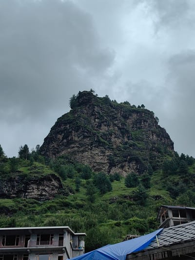 The image captures a dramatic mountainous landscape under an overcast sky. A large, rugged, rocky cliff face dominates the center of the frame, partially covered in dark green vegetation and sparse trees. The cliff rises steeply from a verdant, tree-covered slope. In the foreground, to the right, are parts of two buildings. One is a multi-story structure with exposed concrete and windows. The other, partially visible at the very bottom right, is a tiled roof with a blue tarp covering part of it. The scene appears to be located in a rural or mountainous area, likely Palchan, India, given the context provided. There are no people or discernible actions in the image. The environment is characterized by natural elements and construction.