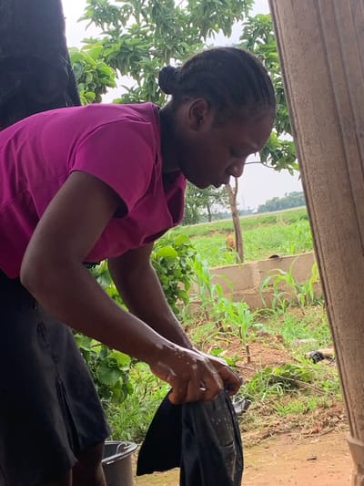 A young Black woman with braided hair styled in two buns is standing and washing dark fabric in a bucket. She is wearing a bright pink t-shirt and a black skirt. Her hands are wet, and she is actively wringing out the fabric. In the background, there is lush green vegetation, including trees and small plants. A rough, textured wall or pillar is visible on the right side of the frame. The ground appears to be dirt and there's a small gray bucket. The environment suggests an outdoor setting in a rural or semi-rural area, possibly within Agban, Nigeria, given the context. The lighting suggests an overcast or diffused daylight.