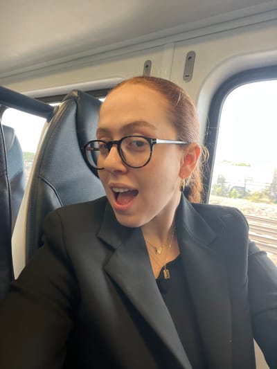 A female subject, wearing a black blazer over a black top and round tortoiseshell-patterned eyeglasses (Tom Ford branded), is depicted in a medium close-up within a transit vehicle. Her red hair is pulled back, and she has a gold hoop earring on her visible right ear. Two layered gold necklaces are visible; one features a butterfly charm, and the other a rectangular pendant with an initial. Her mouth is open, revealing her teeth, as she looks towards the camera. She is seated in a dark grey upholstered seat. Through the window on the right, multiple parallel railway tracks are visible, extending into the distance alongside green foliage and indistinct background structures under an overcast sky. The scene is consistent with a train traveling through an area like Linden, New Jersey, United States.