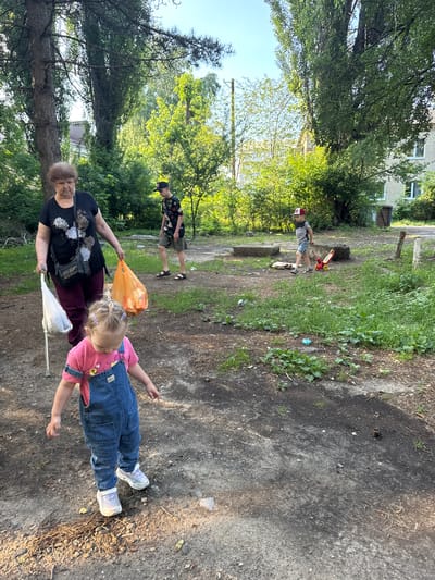 In this outdoor scene, likely in Верхнерусское, Russia, several individuals are present. A young child, wearing denim overalls over a pink shirt and white sneakers, is lying on the ground. An older woman, dressed in a black t-shirt and dark pants, stands nearby, holding an orange plastic bag and a white object, possibly a broom or net. To the left and further back, a young boy in a patterned shirt and shorts, and a younger boy wearing a red cap and grey t-shirt, are also visible, with the younger boy standing near a red scooter.
The setting appears to be a somewhat unkempt outdoor area with patches of grass and dirt paths, possibly a park or a communal yard. Trees with lush green foliage surround the area, suggesting it is daytime, likely late spring or summer. The sunlight filtering through the trees indicates it is not overcast. The overall atmosphere seems relaxed, possibly indicating a casual outing or cleanup activity. No visible text is present in the image.