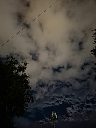 An image of a dark sky with clouds, taken at night in Nalchik, Russia. In the lower center, a telecommunications tower is visible, its antennas illuminated. To the left, the dark silhouette of a tree with dense foliage is prominent. Several power lines stretch diagonally across the frame, obscuring portions of the sky. The lighting suggests an urban environment, possibly with streetlights or building lights illuminating the clouds from below, creating a dramatic, moody atmosphere. There are no people or active events depicted in the image.