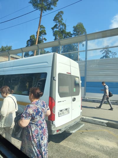 The image depicts a street scene in Zhukovsky, Russia, with a white minibus as the central focus. Two women are standing near the rear of the minibus, one in a cream-colored outfit and the other in a floral dress. A man in a grey t-shirt and pants is walking away from the minibus, looking at his phone. The minibus has a license plate reading "M698CT 750 RUS" and a visible route number "17K". In the background, a sound barrier with reflective panels lines the street, behind which tall pine trees are visible against a bright blue sky. The ground is paved with asphalt and concrete, with yellow markings visible on the asphalt. The lighting suggests it is daytime, likely a sunny day.