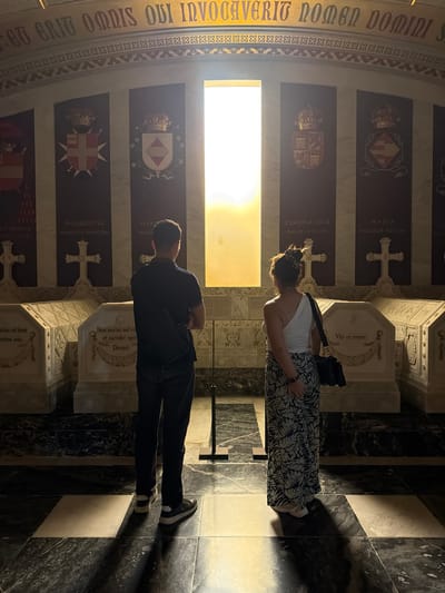Two individuals, a man and a woman, stand facing away from the camera in a grand mausoleum, likely the Royal Pantheon of San Lorenzo de El Escorial, Spain. The man, on the left, wears a dark blue polo shirt and dark trousers, his arms crossed. The woman, on the right, is dressed in a white one-shoulder top and a black and white patterned skirt, carrying a black bag over her shoulder. They are positioned in front of several ornate sarcophagi. Above the sarcophagi are large panels bearing coats of arms and names, illuminated by a bright light source emanating from a large window or opening at the back of the room. The floor is a striking black and white checkered marble pattern, with significant shadow play. The architectural style suggests a historical and possibly royal burial site, consistent with its location within the Monastery of San Lorenzo de El Escorial.