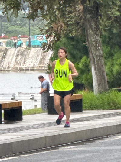 A woman in a neon yellow tank top with the word "RUN" printed in black, and black shorts, is jogging on a paved path. She is wearing pink and blue running shoes. She has dark hair pulled back. Behind her, to the left, a man in a light blue t-shirt and grey pants stands with his head bowed. He is facing away from the camera, looking down towards his feet. In the background, there is a body of water with a concrete retaining wall. Further in the distance, a teal-colored vehicle is parked near some planters with red flowers. Trees and lush green vegetation are visible in the background. Two wooden benches with dark bases are placed on the paved area. The environment appears to be a park or waterfront area, possibly in the city of Xuân Hương - Đà Lạt, Vietnam, given the presence of the lake and park-like setting.