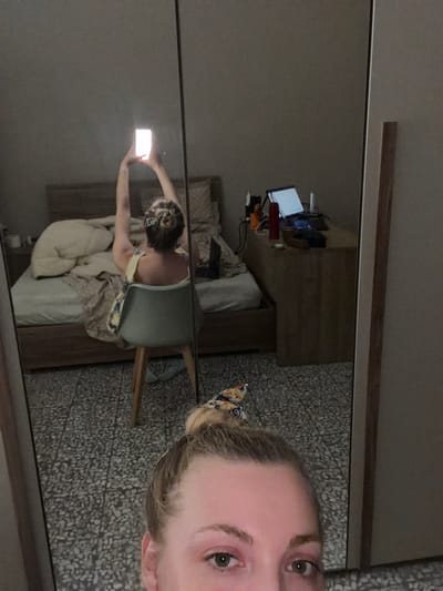 The image shows a reflection in a large mirror of a woman in a bedroom. The woman is sitting in a chair in front of a desk with a laptop. She is holding a phone up to take a selfie. The room appears to be a bedroom with a bed, a nightstand, and a closet. The flooring is tiled. The setting is likely a residential apartment. The image was taken in Catania, Italy.