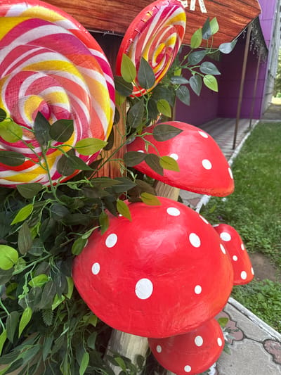 In Pavlodar, Kazakhstan, this image captures an outdoor decorative arrangement featuring oversized, whimsical elements. The foreground is dominated by a collection of red and white spotted mushroom decorations, with the largest positioned prominently in the center. To the left, a large, swirled lollipop decoration in vibrant colors of red, yellow, and white is visible. Artificial green foliage is interspersed throughout the display, adding a touch of artificial nature. In the background, a section of a building with purple walls and a gray, patterned pathway can be seen, suggesting an urban or semi-urban setting. The ground is covered with green grass and stone paving.