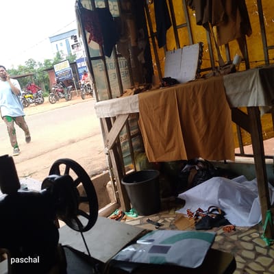 The image depicts a street scene in Nsugpe, Nigeria, captured from inside a tailor's shop. A man is walking on the street while using a mobile phone. Three men on motorcycles are parked nearby. The tailor's shop is cluttered, with fabrics, a bucket, and a sewing machine. A table covered with fabric and other materials is also visible. There are signs visible in the background, one reading "SKY GOLD HOTEL" and another partially visible sign reading "SKYLAND ACADEMY Close To Descent Oil & Gas".