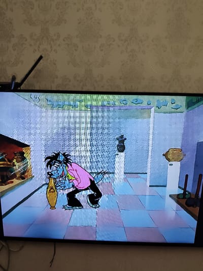 The image displays a television screen showing a scene from an animated cartoon. A blue, anthropomorphic wolf character, wearing a pink shirt and black pants, is depicted in what appears to be a museum or exhibit hall. The character is holding a large, golden amphora. The background includes display pedestals with artifacts, tiled flooring, and patterned wallpaper visible above the screen. The television itself is mounted on a wall with the wallpaper. There is a black antenna or stand protruding from the top left corner of the television. No specific landmarks or elements definitively place the scene in Novorossiysk, Russia.
