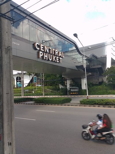 The image captures a street view in Wichit, Thailand, featuring a prominent pedestrian bridge connected to the "Central Phuket" shopping complex. The bridge, made of dark gray material, is adorned with large, illuminated white lettering spelling out "CENTRAL PHUKET" on its side. Above the lettering, the bridge's facade is constructed with large glass panels, reflecting the sky and clouds. A street lamp stands near the bridge, and power lines crisscross the upper portion of the image.

Below the bridge, the environment is lush with greenery, including well-maintained hedges bordering the sidewalk and street. On the road in the foreground, a blurry image of two individuals riding a motorcycle suggests movement and daily activity. The motorcycle is white with red accents. The road itself is asphalt, marked with white lines. In the background, beyond the bridge and greenery, glimpses of buildings and trees are visible, hinting at the urban setting. A small bus stop shelter with yellow seats is situated near the base of the bridge.