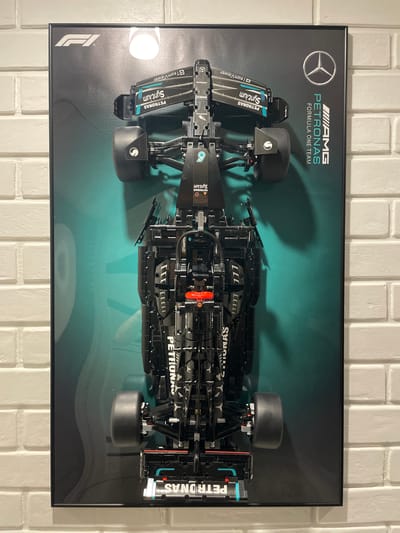 The image shows a framed Lego replica of a Mercedes-AMG Formula One car, mounted on a brick wall. The car is black with teal and white accents, featuring sponsor logos such as PETRONAS, SYNCKING, and TEAMVIEWER. The background of the frame is a gradient from dark grey to teal, with faint circular patterns. The frame itself is black and appears to be hanging on a white brick wall. There is a reflection of a light source on the surface of the frame. The specific location within the city of Sumy, Ukraine, is not determinable from the image alone. There are no people or actions depicted in the image, only the static display of the Lego car.