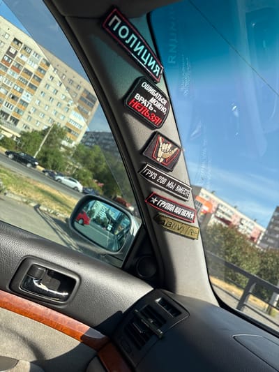 The image shows the interior of a car parked on a street in Biysk, Russia, on a sunny day. The view is through the driver's side window. The car's interior includes a gray leather door panel, a chrome door handle, and a wooden trim piece. A sun visor is visible on the top right of the frame.
Several patches are adhered to the car's A-pillar, which is the pillar between the windshield and the front door. The patches have text and images on them, some of which are in Russian. One patch says "ПОЛИЦИЯ" (Police). Another patch has text that translates to "It's okay to make mistakes, it's forbidden to lie!". Another patch depicts a hand gesture with the "rock on" sign. Two other patches have text that appears to be in Russian, one of which reads "ГРУЗ 200 МЫ ВМЕСТЕ" (Cargo 200 we are together) and another that appears to be "ГРУППА ВАГНЕРА" (Wagner Group). The bottom patch is olive green and has the text "AB(IV)R+".
Outside the car, apartment buildings and trees are visible. The sky is blue and clear. Cars are driving on a road in the background. A side mirror is also visible, reflecting the outside environment.