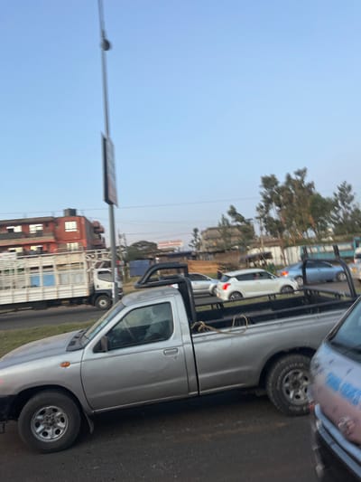 The scene depicts a street in Biashara ward, Kenya, featuring multiple vehicles on a paved road under a clear, light blue sky.

In the foreground, a silver, two-door pickup truck, consistent with a Nissan D21 or Hardbody model, is parked or moving slowly. It is equipped with a black, tubular roll bar or bed rack over its cargo area. A driver is vaguely visible inside the cabin. To the immediate right, the front portion of a gray van or minibus is partially visible.

In the midground, a two-lane road shows ongoing traffic. A white flatbed truck with a white and blue cage-like body on its bed is visible on the left lane, moving from left to right. Other vehicles in the traffic flow include a white Suzuki Swift hatchback, a silver sedan, and a blue sedan, positioned behind the prominent silver pickup truck.

The background features a reddish-brown, two-story building with white-framed windows on the left. A tall, gray pole with an unidentifiable rectangular sign is present in the center. Clusters of green trees are visible in the mid-distance and on the right side of the frame. Natural daylight illuminates the scene, suggesting daytime or late afternoon. No specific interactions between individuals are discernible, only vehicular presence on a thoroughfare.