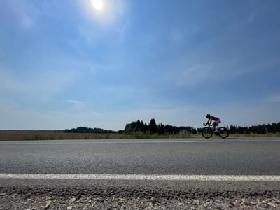 A person in a red and white cycling outfit rides a road bicycle along a paved road. The cyclist is wearing a helmet and appears to be in motion. The road has a white line marking its edge. In the background, a line of trees forms a dark band against a pale sky. The sun is visible in the upper left corner, partially obscured by wispy clouds. The foreground shows the rough texture of the road's shoulder, composed of small stones and gravel. The image depicts an outdoor scene under a clear, bright sky.
