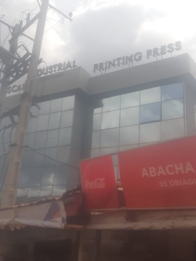 The image shows a building with prominent signage indicating "INDUSTRIAL PRINTING PRESS" and "SC." Another sign reads "ABACHA" and "55 OBIAGU." A Coca-Cola logo is visible on a red banner. The building has a modern facade with large glass windows. In the foreground, a utility pole with electrical wires is visible, and a corrugated roof structure partially obscures the lower part of the building. The sky is overcast. The scene is in Enugu, Nigeria.