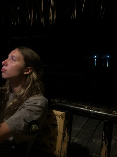 A young woman with light brown hair, wearing a grey t-shirt with a circular patch on the sleeve, is seen from the chest up, looking upwards to her left. She is sitting on a bamboo chair in what appears to be an outdoor dining area. The background is dark, with hints of natural thatch overhead and a dimly lit body of water with lights reflected on it in the distance. The scene suggests a nighttime setting, possibly at a restaurant or resort in Kertasari, Indonesia, a location known for its coastal views and natural beauty.