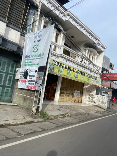 The image shows a storefront in Kecamatan Margahayu, Indonesia, located at coordinates -6.976 latitude and 107.565 longitude. The storefront has a large sign that reads "MEUBEL SUMBER JEPARA" in yellow letters on a green background. Below the sign, the storefront is covered with a wooden door with graffiti painted on it in black and white. The storefront is located on the corner of a street, with a sidewalk in front of it. To the left of the storefront is a sign that reads "OPTIK KRIDA" in red letters on a white background, with a smaller green sign underneath that says "MELAYANI". Below this sign is another sign that reads "BRIS Kesehatan" in black letters on a white background. The storefront is located in an urban setting with numerous powerlines running overhead. There is one person visible in the image, a man wearing a red shirt, who is walking on the sidewalk to the right of the storefront. The street is paved with asphalt. The weather is sunny and clear. The image appears to have been taken at midday.