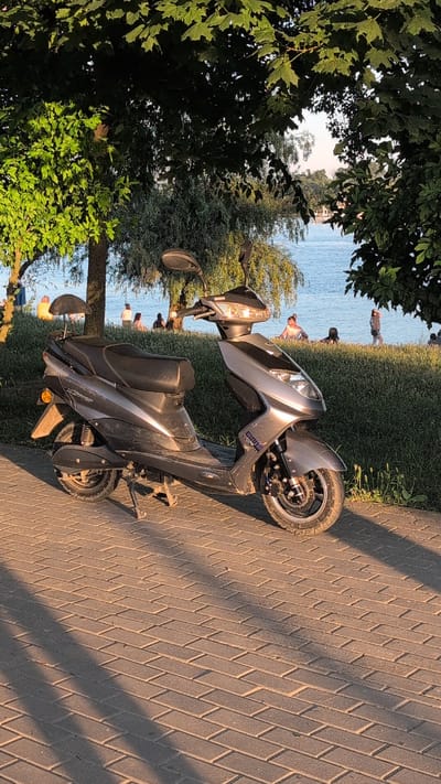 A gray electric scooter is parked on a brick-paved walkway. The scooter is in the foreground, facing left, with its kickstand down. Behind the scooter, a grassy embankment slopes down to a body of water. Several people are visible on the embankment and in the shallow water near the shore. Trees with green leaves are present on the left and right sides of the image, casting shadows on the walkway. The scene appears to be a public park or waterfront area, possibly in Izmail, Ukraine, given the provided context. The lighting suggests it is late afternoon or early evening, with warm sunlight.