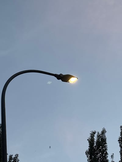 A street lamp with a bright yellow light is illuminated against a cloudy, grey sky. The lamp is a modern design with a dark, curved arm extending from the right side of the frame, swooping upwards to hold the light fixture. In the upper right corner of the image, the dark silhouettes of tree branches with dense foliage are visible against the sky. The overall lighting suggests it could be dusk or an overcast day. There are two small, indistinct smudges or marks in the sky, possibly artifacts or distant objects. No people or text are visible in the image. The location context provided is Coslada, Spain.