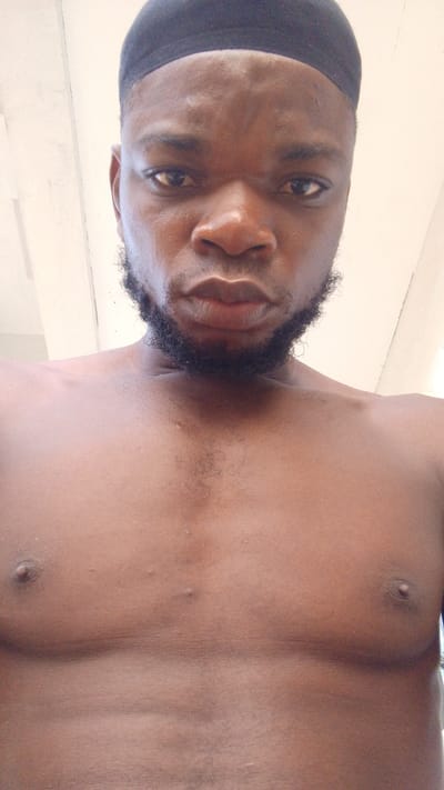 A close-up, eye-level shot of a Black man with a dark beard and a black cap. He is shirtless, revealing his chest. The location is indicated as Enugwu-Ukwu, Nigeria, although this is not evident from the image itself. The background appears to be a plain, light-colored wall or ceiling. The man is looking directly at the camera.
