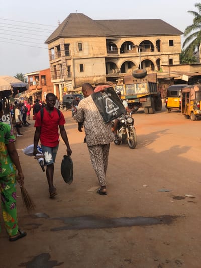 This image captures a daytime street scene in Neni, Nigeria. The foreground features an unpaved, reddish-brown dirt road with scattered debris and dark stains. Sunlight casts clear shadows, suggesting mid-morning or afternoon.
Three individuals are prominent in the foreground:
1. On the far left, a person is partially visible, wearing green trousers with a vibrant geometric pattern and dark sandals, holding a small, natural-fiber broom.
2. In the center-left, a young male walks towards the viewer, looking slightly right. He wears a red short-sleeved t-shirt, distressed denim shorts with a white leaf pattern, and dark flip-flops. He carries a white bundled item in his left hand and a dark, possibly fabric bag in his right.
3. In the center-right, a male walks away from the viewer, dressed in a patterned, light brown or beige two-piece traditional Nigerian outfit and dark sandals. He carries a black plastic bag with "Eco Friendly 05 PP" and a recycling symbol printed in white, slung over his left shoulder.
In the midground and background, street activity is evident. To the left, several indistinct figures are gathered under market stalls with blue tarpaulin awnings. Vehicles are present in the midground and background:
* A blue and yellow dump truck with a tire resting on its bed and the number "56" on its side is visible to the right of center.
* A dark-colored motorcycle with a rider is visible behind the patterned-clothed man.
* Two yellow motorized tricycles, known as Keke Napeps, are parked or moving on the far right.
* A dark van or SUV is visible further in the background on the left.
The background is dominated by buildings. A prominent, light-colored, possibly unfinished three-story building with a brown hip roof stands in the center-right. It features multiple arched openings on the upper floors and rectangular windows. Smaller, reddish-brown buildings are visible on the left. A palm tree is partially visible on the far right. Overhead power lines traverse the upper portion of the frame.