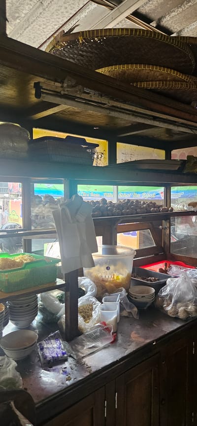 This is an outdoor food stall in Bekasi, Indonesia. The stall has a wooden structure with shelves displaying various food items and cooking utensils. On the top shelf, there are woven bamboo trays. Below that, a blue basket holds some items. The middle shelf showcases a large quantity of round, brown food items, likely meatballs, arranged on a rack. In front of this, a clear plastic container is filled with light-colored, crispy snacks.
The lower counter area is cluttered with food supplies. A green basket contains what appears to be noodles. Several stacks of white bowls are visible, along with plastic bags filled with dried noodles, and small containers of what might be spices or condiments. There are also some larger plastic bags containing round food items, possibly more meatballs. A white cloth hangs down from the structure, obscuring some of the items. The environment appears to be somewhat informal, with evidence of daily use and preparation for serving food. The lighting suggests it is daytime, possibly with sunlight filtering through.
