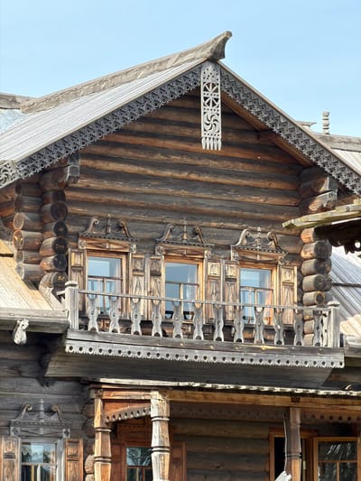 The image shows the exterior of a traditional wooden log house in Nevskii Parkleskhoz, Russia. The house features ornate wooden carvings around the windows, balconies, and eaves, characteristic of Russian folk architecture. The main focus is on the upper part of the house, including a prominent roof with decorative trim and a balcony with carved railings. The logs used in the construction are visible, showing the natural texture and color of the wood. The sky above is clear and pale blue. There are no people or animals visible in the image, and no specific actions or interactions are depicted. The environment appears to be outdoors, likely in a rural or park-like setting.