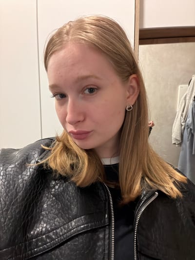 A young Caucasian woman with blonde hair is facing the camera, wearing a black leather jacket. She has fair skin and a stud earring. The background includes a light-colored wall, a white door, and a garment rack with clothes. The location is unknown, as there is nothing specific to indicate it is in Rossiyskiy, Russia.